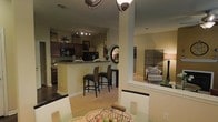 This single-story residence is designed with an open-concept layout that seamlessly connects multiple living areas. The kitchen is outfitted with dark wood cabinetry reaching toward the ceiling, dark countertops, and black appliances, including a built-in microwave. Track lighting illuminates the workspace effectively. A raised breakfast bar with dark granite-style countertops extends from the kitchen, providing casual seating for two with wicker-style bar stools — an ideal spot for quick meals or entertaining.

Adjacent to the kitchen, a dining area is anchored by a round glass-top table with a decorative centerpiece, lit by a ceiling fan fixture overhead. The open floor plan transitions naturally into a sitting area or secondary living space, visible through a wide architectural opening framed by a structural column. That space features a cushioned armchair with decorative pillows, a tall floor lamp, and a latticed decorative screen, contributing to the warm, inviting ambiance. Neutral beige walls and light carpeting in the living areas contrast with the darker tones of the kitchen, creating a balanced and cohesive aesthetic throughout the home.