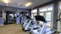 This fitness center offers a comprehensive workout environment designed to accommodate a range of exercise routines. The space is outfitted with a row of Matrix brand treadmills and elliptical machines positioned to face large, multi-pane windows that allow abundant natural light to fill the room. The windows are framed with cream-colored trim and are complemented by glass-paneled doors that further enhance the bright, open atmosphere.

The walls are finished in a deep blue-gray tone, lending the space a modern and focused ambiance. Full-length mirrors line one side of the room, visually expanding the area while allowing users to monitor their form during workouts. A dedicated free weights section is visible in the mirrored reflection, complete with a dumbbell rack and additional strength training equipment.

Overhead, ceiling fans provide consistent air circulation throughout the space, supporting comfort during intense exercise sessions. The flooring features a textured, patterned carpet. Recessed and panel lighting supplements the natural light, ensuring the space remains well-lit at all hours. Overall, the fitness center is thoughtfully arranged to balance cardio equipment, strength training, and open movement space in a comfortable, well-lit setting.