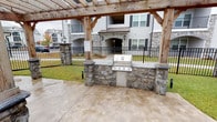 This single-story community clubhouse combines stylish interior spaces with thoughtfully designed outdoor amenities. Inside, the main lounge area features high ceilings adorned with crown molding, light hardwood flooring, and soft gray walls that create a calm, welcoming atmosphere. A wall-mounted flat-screen TV, comfortable seating including a blue sofa and accent chairs, and a media credenza make this a functional gathering space. Decorative framed artwork and vibrant patterned drapes add personality throughout.

Adjacent to the lounge, a bright workspace area is framed by expansive floor-to-ceiling black-framed windows that flood the room with natural light and offer direct views of the outdoor pool and pergola. A built-in work counter with computer stations and a glass-top bistro table with gold accent chairs provide both productivity and social options in this airy setting.

Outside, a substantial rustic timber pergola with stone column bases anchors the outdoor entertaining area. A built-in stainless steel gas grill set into a stone countertop and base serves as the centerpiece of the outdoor kitchen. The concrete patio connects seamlessly to the surrounding green lawn, iron fencing, and views of the broader residential community, making it an ideal space for outdoor gatherings and leisure.