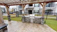 This single-story community clubhouse combines stylish interior spaces with thoughtfully designed outdoor amenities. Inside, the main lounge area features high ceilings adorned with crown molding, light hardwood flooring, and soft gray walls that create a calm, welcoming atmosphere. A wall-mounted flat-screen TV, comfortable seating including a blue sofa and accent chairs, and a media credenza make this a functional gathering space. Decorative framed artwork and vibrant patterned drapes add personality throughout.

Adjacent to the lounge, a bright workspace area is framed by expansive floor-to-ceiling black-framed windows that flood the room with natural light and offer direct views of the outdoor pool and pergola. A built-in work counter with computer stations and a glass-top bistro table with gold accent chairs provide both productivity and social options in this airy setting.

Outside, a substantial rustic timber pergola with stone column bases anchors the outdoor entertaining area. A built-in stainless steel gas grill set into a stone countertop and base serves as the centerpiece of the outdoor kitchen. The concrete patio connects seamlessly to the surrounding green lawn, iron fencing, and views of the broader residential community, making it an ideal space for outdoor gatherings and leisure.