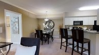This single-floor residence offers a thoughtfully designed open-concept layout that seamlessly connects the living, dining, and kitchen areas. The dining space is anchored by a sleek dark-wood table paired with matching upholstered chairs, centered beneath an elegant pendant light. A large decorative round mirror mounted on the warm-toned wall adds depth and sophistication to the room, while tall ornamental grasses in floor vases contribute a refined, contemporary aesthetic.

Adjacent to the dining area, the kitchen is outfitted with crisp white upper cabinetry, dark granite countertops, and black appliances, creating a striking contrast that feels both modern and functional. A raised breakfast bar with tall bar-height chairs provides an additional casual dining option and further connects the kitchen to the main living space.

Soft, neutral-toned carpet flows throughout the living and dining areas, contributing to the overall warmth of the home. Crown molding along the ceiling adds an elegant architectural detail, enhancing the polished character of the interior. The living area, partially visible in the foreground, features a dark upholstered sofa that complements the cohesive design palette. Overall, the space presents a clean, contemporary style with a welcoming and comfortable ambiance.