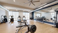This modern fitness center offers a generously sized, single-level open floor plan designed to accommodate a full range of workout activities. The space features light wood-tone flooring throughout, crisp white walls accented by bold navy blue focal walls, and exposed industrial-style ductwork that adds a contemporary edge to the overall aesthetic. Recessed linear LED lighting and large ceiling fans ensure the environment remains well-lit and comfortable during use.

Large floor-to-ceiling windows line multiple walls, flooding the interior with abundant natural light and creating an open, airy atmosphere. Full-length mirrors are strategically positioned along one wall, enhancing the sense of space and allowing users to monitor their form during exercise.

The equipment layout is thoughtfully organized, with dedicated zones for various training modalities. Cardio equipment — including treadmills, ellipticals, rowing machines, and stationary bikes — occupies one section, while a separate strength training area houses cable machines, multi-function press stations, and adjustable benches. A free weight zone features a full rack of dumbbells, and a functional training corner includes a heavy bag and boxing equipment. A glass-paned entry door provides easy access, completing this comprehensive and well-appointed fitness facility.