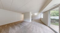 This single-floor residence is designed with a practical, open layout that maximizes the available living space. The main living area is generously proportioned, featuring neutral-toned carpet underfoot and soft beige walls throughout. A large sliding glass door floods the space with natural light and provides views of the surrounding greenery, while an additional window offers supplemental brightness. Structural ceiling beams add subtle architectural character to the room.

The layout includes a distinct open area that flows seamlessly, with a central dividing wall adding definition between living zones. The kitchen is situated off the main living space and features dark espresso-toned cabinetry, light countertops, and durable laminate flooring. White appliances, including a refrigerator, dishwasher, and range, are provided.

The bedroom is a well-sized, carpeted room equipped with a standard closet with paneled doors. The bathroom includes a wide vanity with a white countertop, under-sink cabinetry, a mirrored medicine cabinet, and a separate glass-door shower enclosure with a private water closet. The overall ambiance is clean, neutral, and move-in ready.