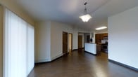 This single-floor space presents an open-concept layout that seamlessly connects the living area to the kitchen. Dark hardwood flooring runs throughout, providing a warm, cohesive foundation. The main living area features neutral cream-colored walls and a decorative pendant light with a frosted glass shade that serves as a central focal point overhead. A baseboard heating unit lines one wall, offering practical climate control.

The kitchen is positioned toward the rear of the space and is equipped with dark wood cabinetry, a stainless steel gas range, and a microwave. A white kitchen island or peninsula creates a natural division between the cooking and living areas. Recessed fluorescent lighting illuminates the kitchen workspace, complementing the darker cabinetry tones. A washer unit is also visible near the kitchen, indicating in-unit laundry access.

Multiple doorways with dark wood trim lead to additional rooms, suggesting a functional layout that includes separate private spaces. The open corridor view hints at a bathroom and at least one additional room. Overall, the space maintains a straightforward, practical design with classic finishes throughout.
