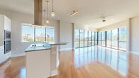 This single-floor residence is defined by its striking floor-to-ceiling glass walls that frame panoramic city skyline views, flooding the open-concept living and dining areas with abundant natural light. Warm hardwood flooring extends throughout the main living spaces, complementing the contemporary design aesthetic.

The kitchen is thoughtfully designed with a large central island featuring an induction cooktop and an overhead stainless steel range hood, suspended pendant lighting, crisp white cabinetry, a tile backsplash, and stainless steel appliances including wall ovens and a refrigerator. The kitchen flows seamlessly into the expansive living area, which is anchored by corner floor-to-ceiling windows and sliding glass doors that open to outdoor access.

The bedrooms are carpeted and bathed in natural light through city-facing windows, offering generous proportions and a calm, neutral palette. One bathroom features a soaking tub with tile surround and a separate glass-enclosed shower, while another includes a glass walk-in shower and direct access to a spacious walk-in closet outfitted with multiple shelving tiers and hanging rods. Throughout, the residence maintains a clean, modern character with neutral tones and refined finishes.