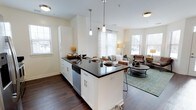 This single-floor, two-bedroom, two-bathroom residence showcases a thoughtfully designed open-concept layout that seamlessly connects the kitchen, dining, and living areas. The kitchen serves as the centerpiece of the space, featuring a large island with a dark granite countertop that provides both prep space and casual seating. White shaker-style cabinetry, stainless steel appliances including a dishwasher, and a deep sink contribute to a clean, contemporary aesthetic. Pendant lighting with polished nickel fixtures hangs above the island, adding a refined touch. Rich, dark hardwood flooring flows throughout, creating a warm contrast against the light-toned walls. The adjoining living area is bright and airy, benefiting from multiple windows dressed with horizontal blinds that invite generous natural light. The open floor plan creates a sense of spaciousness, making the common areas feel connected and welcoming. Overall, the residence strikes a balance between modern functionality and comfortable, everyday living, making it an inviting and practical space.
