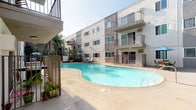 This outdoor communal space is centered around a generously sized freeform swimming pool with clear, blue-green water, framed by a wide concrete pool deck ideal for lounging. The surrounding multi-story building features a classic stucco exterior in neutral grey and white tones, with sliding windows and private balconies overlooking the pool area. The open courtyard layout allows abundant natural sunlight to fill the space throughout the day, creating a bright and inviting atmosphere. Poolside amenities include cushioned lounge chairs positioned for sunbathing, as well as a dedicated outdoor dining area furnished with a wooden table, chairs with green cushions, and a large blue umbrella for shade. Wrought iron fencing encloses portions of the deck, adding a sense of structure and security to the space. Tropical and ornamental plantings, including potted palms and climbing greenery along the perimeter fencing, introduce a lush, resort-inspired feel. The overall ambiance is relaxed and California-casual, designed to encourage outdoor leisure and social gathering in a sun-drenched setting.