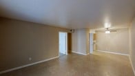 This single-floor space is characterized by a clean, open layout designed for functional everyday living. The main living area is generously proportioned, offering a bright and airy feel enhanced by smooth white ceilings and warm neutral-toned walls that create a calm, welcoming atmosphere. The flooring throughout features light-toned vinyl tile, providing a practical and easy-to-maintain surface that complements the understated color palette. White baseboards and door trim add a crisp, finished contrast to the warm beige walls. The living area connects seamlessly to additional rooms via interior doorways, with one door leading to a secondary room or hallway and another providing a direct sightline into what appears to be a kitchen, where wood-toned cabinetry is visible. The kitchen area is positioned at the far end of the space, offering a natural separation from the living area while maintaining an open flow. Ceiling-mounted HVAC vents are integrated into the layout, ensuring year-round climate comfort. The overall design is simple and well-maintained, with a neutral, move-in-ready aesthetic that offers a versatile canvas for a variety of furnishing and décor styles.