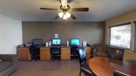 This business center is a practical and well-equipped space designed to support productivity and connectivity. The room features a built-in wooden workstation counter along one wall, housing multiple desktop computer terminals, each with its own monitor and ergonomic rolling chair — providing ample workspace for several users simultaneously. The warm oak cabinetry beneath the counter adds a welcoming touch to the otherwise functional setup. Informational materials and printed resources are displayed at each station for easy reference. A ceiling fan with a light fixture is centrally mounted overhead, ensuring comfortable air circulation throughout the space. The room is finished with neutral taupe-toned walls and low-pile carpeting, contributing to a quiet, focused atmosphere. To the side, a round wooden table accompanied by a leather chair offers an additional informal seating and working area. A comfortable upholstered armchair is also present, providing a more relaxed option for visitors. The overall layout is efficient and user-friendly, making this space a convenient resource for anyone in need of computer access, printing services, or a quiet place to work.