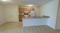 This single-floor space is designed with an open layout that connects the kitchen and living area, creating a functional and cohesive environment. The kitchen is equipped with medium-toned oak cabinetry offering ample storage, dark-speckled laminate countertops, and white appliances including a gas range and a full-sized refrigerator. A built-in peninsula extends from the kitchen, providing additional counter space and a natural divider between the kitchen and the adjacent living area. The peninsula also features a sink, making it a practical workspace. The kitchen is finished with neutral tile flooring, which transitions into carpeting in the living area, adding a layer of warmth and comfort underfoot. The walls are painted in a light, neutral tone throughout, contributing to a clean and airy feel. Recessed ventilation and a flush-mount ceiling light illuminate the kitchen. Two doors are visible — one likely leading to a closet and another to an additional room — suggesting a practical floor plan. The textured ceiling adds a subtle traditional detail to the overall aesthetic.