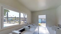 This single-story laundry facility offers a clean and functional environment designed to serve its users efficiently. The room is finished with smooth, neutral-toned walls and a bright white ceiling that contributes to an open and airy atmosphere. A large picture window along one wall floods the space with abundant natural light, offering views of the surrounding outdoor area, including mature trees, a parking area, and neighboring buildings. The open doorway at the far end of the room provides additional natural light and a direct view to the exterior courtyard, where landscaping and a blue-accented building are visible, enhancing the sense of connection to the outdoors. The facility is equipped with multiple top-loading washing machines and dryers arranged in rows, providing ample capacity for residents. The machines are white and neatly aligned, contributing to the room's tidy and organized appearance. The flooring is light-toned and easy to maintain, well-suited for the functional demands of the space. Overall, the laundry room balances practicality with a surprisingly pleasant and well-lit environment, making it a convenient and comfortable shared amenity.