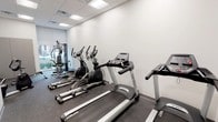 This fitness center is designed to offer a functional and efficient workout environment within a clean, modern aesthetic. The space features white walls, a drop ceiling with recessed panel lighting, and durable dark speckled rubber flooring throughout, providing both practicality and a polished appearance.

The main workout area houses a row of Spirit-brand treadmills and elliptical trainers positioned along one wall, offering ample space between machines for comfortable use. A multi-function cable and weight machine is situated toward the windowed end of the room, where natural light filters in through a street-facing window, adding brightness to the space.

A secondary area, partially separated by a structural column, provides additional room for flexible training. This section includes a recumbent bike, a rowing machine, and a balance trainer, accommodating a variety of low-impact and cross-training exercises. A wall-mounted thermostat, a small waste bin, and a sanitation station reflect thoughtful attention to user comfort and hygiene.

Glass-panel exit doors connect to an adjacent corridor, ensuring easy access. Overall, the fitness center maintains a clean, no-frills atmosphere well-suited for daily exercise routines.