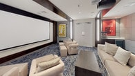This elegantly appointed private screening room is designed to deliver a premium cinematic experience. At the heart of the space is a large, wall-mounted projection screen paired with a ceiling-mounted projector, ensuring crisp, high-quality viewing from every seat. The room is furnished with generously proportioned cream-toned sofas, lounge chairs, and ottomans, all arranged across a striking blue and ivory patterned carpet that adds a layer of visual texture and warmth underfoot. Acoustic fabric wall panels in a neutral grey line the perimeter, providing both sound insulation and a refined aesthetic. Subtle vertical LED light strips are integrated into the walls, casting a soft, ambient glow that complements the recessed ceiling lighting. A dark wood structural column anchors the center of the room, contributing architectural character while dividing the seating into distinct zones. Broadway show posters — including "The Wiz" and "West Side Story" — are displayed throughout, lending a playful, cultural touch to the sophisticated interior. A secondary lounge area with a sofa and coffee table sits toward the rear, ideal for pre-screening gatherings. The space is accessed through sleek double doors flanked by illuminated panels, completing a polished and immersive entertainment environment.