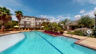Located at 9611 Grant Rd in Houston, Texas, this residential community amenity space is designed to offer a resort-style outdoor living experience. The centerpiece of the community is a generously sized swimming pool with crystal-clear blue water, flanked by vibrant tropical landscaping including tall palm trees, colorful flowering plants, and manicured hedges. The pool deck is equipped with bright orange chaise lounges and shaded umbrella seating, providing ample space for sunbathing and relaxation under the Texas sky.

Adjacent to the pool is a covered outdoor lounge area that serves as a comfortable gathering space. This area features bold orange modular seating accented with striped throw pillows, brick accent columns, and paver flooring that adds a warm, textured aesthetic. Large floor-to-ceiling glass panels open the lounge to the surrounding grounds, blending indoor comfort with the outdoors seamlessly. Recessed lighting and a decorative chandelier visible through the glass enhance the ambiance during evening hours.

The multi-story residential buildings surrounding the amenity space are finished in a classic gray and blue palette with white trim, bay windows, and pitched rooflines. Lush greenery and flowering plants are integrated throughout the grounds, creating a well-maintained, park-like setting that enhances the overall atmosphere of the community.