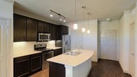 Located in North Richland Hills, TX, this single-story home showcases a thoughtfully designed interior with modern finishes throughout. The kitchen serves as a central highlight, featuring rich dark cabinetry that creates a striking contrast against the light granite countertops. A curved kitchen island with a built-in stainless steel sink provides additional workspace and serves as a natural gathering point. Pendant lighting hangs above the island, adding warm, focused illumination, while track lighting overhead ensures the entire kitchen area is well-lit. The kitchen is fully equipped with stainless steel appliances, including a French door refrigerator, electric range, and built-in microwave. Dark hardwood flooring runs throughout, adding warmth and continuity to the space. The layout flows naturally toward adjacent areas, with clean white walls and panel-style doors contributing to a crisp, contemporary aesthetic. The overall ambiance of the home balances functionality with style, making it a practical yet visually appealing living space suited for everyday comfort.