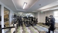 This fitness center offers a comprehensive workout environment housed on a single floor. The space is outfitted with a diverse selection of exercise equipment, including multiple treadmills, stationary bikes, elliptical machines, and a recumbent bike, catering to a wide range of cardio and low-impact training preferences. Additional strength and conditioning equipment is positioned along the sides of the room, maximizing usable floor space.

The interior is finished with a distinctive geometric-patterned carpet in shades of green, blue, and gray, adding a modern, energetic aesthetic to the environment. Overhead, recessed troffers and flush-mounted fluorescent light fixtures provide consistent, bright illumination throughout the space. The ceiling features subtle coffered detailing, lending a polished architectural touch.

Neutral gray walls and white doors contribute to a clean, uncluttered atmosphere. Mirrors are positioned to visually expand the space and allow users to monitor their form. The layout is open and organized, enabling easy navigation between equipment stations. Overall, the fitness center presents a functional and contemporary setting well-suited for daily exercise routines.