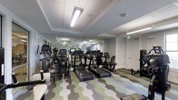 This fitness center offers a comprehensive workout environment housed on a single floor. The space is outfitted with a diverse selection of exercise equipment, including multiple treadmills, stationary bikes, elliptical machines, and a recumbent bike, catering to a wide range of cardio and low-impact training preferences. Additional strength and conditioning equipment is positioned along the sides of the room, maximizing usable floor space.

The interior is finished with a distinctive geometric-patterned carpet in shades of green, blue, and gray, adding a modern, energetic aesthetic to the environment. Overhead, recessed troffers and flush-mounted fluorescent light fixtures provide consistent, bright illumination throughout the space. The ceiling features subtle coffered detailing, lending a polished architectural touch.

Neutral gray walls and white doors contribute to a clean, uncluttered atmosphere. Mirrors are positioned to visually expand the space and allow users to monitor their form. The layout is open and organized, enabling easy navigation between equipment stations. Overall, the fitness center presents a functional and contemporary setting well-suited for daily exercise routines.