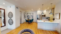 This thoughtfully designed single-floor residence offers a seamless blend of modern style and functional living. The open-concept main living area is anchored by a sleek kitchen featuring white shaker cabinetry, white quartz countertops, a subway tile backsplash, stainless steel appliances including a French door refrigerator and electric range, and a central island with pendant lighting. Warm bamboo hardwood flooring flows throughout, connecting the kitchen to a dining area accented by a striking round wood table, teal velvet chairs, and a bold triptych of abstract wall art. The primary bedroom is a calm retreat with plush carpeting, a ceiling fan, window blinds with drape panels, and direct access to a walk-in closet. The en-suite bathroom impresses with a wide illuminated vanity mirror, crisp white countertops, and a glass-enclosed walk-in shower. A second bedroom offers equally comfortable accommodations, while the second bathroom provides a full tub-shower combination, large-format tile flooring, and a well-appointed vanity. Recessed lighting throughout the space ensures a bright, welcoming ambiance, and the clean white walls provide a neutral backdrop that complements the contemporary décor.