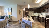 This single-floor residence showcases a thoughtfully designed open-concept layout that seamlessly connects the living, dining, and kitchen areas into one cohesive space. The living area provides a comfortable setting with neutral-toned walls and warm wood-style flooring that extends throughout the unit, creating a consistent and inviting atmosphere.

The kitchen is a standout feature, outfitted with dark espresso cabinetry that reaches toward the ceiling, offering generous storage. A mosaic tile backsplash in muted greens and grays adds visual texture behind the range and countertops. Stainless steel appliances, including a refrigerator, microwave, and dishwasher, provide a polished, modern aesthetic. A curved white breakfast bar island serves as both a functional food preparation surface and a casual dining space, accompanied by contemporary bar seating.

Pendant lighting in elongated cylindrical forms hangs above the island, complementing the overall flush ceiling fixtures to provide layered illumination throughout the space. The open sightlines between the kitchen and living area enhance the sense of spaciousness, making the layout feel airy and well-connected. The combination of dark cabinetry, light countertops, and wood flooring gives the space a balanced, contemporary character.