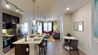 Located at 2323 Wolf Ranch Pkwy in Georgetown, Texas, this single-floor residence offers a thoughtfully designed open-concept layout that seamlessly connects the kitchen, dining, and living areas. The kitchen serves as the heart of the home, featuring a peninsula with bar-height seating, pendant lighting with sleek cylindrical fixtures, a deep undermount sink, and stainless steel appliances. Light-toned countertops contrast elegantly with dark cabinetry, creating a contemporary aesthetic. The open layout flows naturally into the dining area, which accommodates a round bistro-style table and chairs — an inviting spot for casual meals. The living area is warm and welcoming, anchored by a ceiling fan, light hardwood-style flooring, and an accent wall with decorative wallpaper. Large windows dressed with curtains allow ample natural light to fill the space, enhancing the bright and airy ambiance throughout. Comfortable seating options, including a sectional sofa and accent chairs, complete the living area. The consistent hardwood-style flooring throughout ties the open floor plan together, lending the space a cohesive and polished look. Overall, this residence combines functional design with modern finishes to create an comfortable and stylish living environment.
