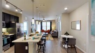 Located at 2323 Wolf Ranch Pkwy in Georgetown, Texas, this single-floor residence offers a thoughtfully designed open-concept layout that seamlessly connects the kitchen, dining, and living areas. The kitchen serves as the heart of the home, featuring a peninsula with bar-height seating, pendant lighting with sleek cylindrical fixtures, a deep undermount sink, and stainless steel appliances. Light-toned countertops contrast elegantly with dark cabinetry, creating a contemporary aesthetic. The open layout flows naturally into the dining area, which accommodates a round bistro-style table and chairs — an inviting spot for casual meals. The living area is warm and welcoming, anchored by a ceiling fan, light hardwood-style flooring, and an accent wall with decorative wallpaper. Large windows dressed with curtains allow ample natural light to fill the space, enhancing the bright and airy ambiance throughout. Comfortable seating options, including a sectional sofa and accent chairs, complete the living area. The consistent hardwood-style flooring throughout ties the open floor plan together, lending the space a cohesive and polished look. Overall, this residence combines functional design with modern finishes to create an comfortable and stylish living environment.