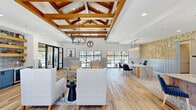 This thoughtfully designed single-floor community space blends modern farmhouse aesthetics with functional, open-plan living. The standout architectural feature is the dramatic coffered ceiling, adorned with rich, warm-toned wood beams and complemented by a statement geometric pendant chandelier, creating an impressive sense of vertical volume throughout the main gathering area. Light-toned wood-look flooring runs continuously across the space, reinforcing its clean, cohesive design.

The open layout seamlessly integrates a comfortable lounge area, a casual dining zone, and a functional reception or workspace. A built-in bar area features blue cabinetry with a beverage cooler, gray tile backsplash, and open natural wood floating shelves, adding a refined yet relaxed character. A dedicated private conference or office room is enclosed by sleek black-framed glass doors, offering a professional workspace within the larger open environment.

A striking natural stone accent wall in warm limestone tones anchors the reception desk area, where pendant lights illuminate a spacious work surface flanked by blue upholstered chairs. Floor-to-ceiling black-framed windows and glass doors flood the interior with natural light and offer unobstructed views of an outdoor pool and surrounding community. The entry corridor features gray shiplap wainscoting, continuing the modern farmhouse theme throughout.