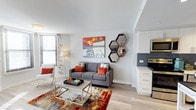 This well-appointed single-floor space showcases a thoughtfully designed open-concept layout that seamlessly connects the living area and kitchen. The living space features light hardwood-style flooring that runs throughout, contributing to a bright and airy atmosphere. Large windows dressed with horizontal blinds allow ample natural light to fill the room, enhancing the clean, modern aesthetic.

The living area is anchored by a contemporary gray sofa accented with rust-orange throw pillows, paired with a sleek glass-and-metal coffee table. A vibrant, multicolored area rug adds warmth and character underfoot, while decorative elements — including a bold, colorful wall painting and a cluster of hexagonal mirrors — lend a curated, artistic touch to the space.

The kitchen, visible from the living area, features crisp white cabinetry, dark slate-toned tile backsplash, and dark countertops, creating a striking contrast that feels both modern and functional. Stainless steel appliances complete the kitchen's polished look.

The space benefits from smooth white ceilings and neutral wall tones, providing a versatile backdrop that balances the vibrant decor. Overall, the unit offers a cohesive blend of contemporary design and comfortable living in a compact, efficiently organized floor plan.