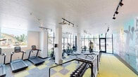 Located at 245 S Orchard St in Boise, Idaho, this fitness center offers a thoughtfully designed, single-floor exercise space that provides a comprehensive workout environment. The gym is divided into distinct functional zones, seamlessly connected by a split-level layout with a metal-railed staircase and glass partitions that maintain an open, airy feel throughout.

The cardio area features a row of treadmills positioned beneath a bright window, complemented by an elliptical trainer and additional cardio equipment. Large floor-to-ceiling windows along the street-facing wall flood the space with abundant natural light while offering views of the surrounding Boise streetscape. Track lighting runs throughout the ceiling, ensuring consistent illumination across all workout zones.

The strength training area is well-appointed with a multi-station cable machine, adjustable benches, and a full rack of dumbbells. Flooring transitions between patterned carpet and bold yellow exercise tiles, clearly delineating workout zones while adding a dynamic visual element to the space.

A standout design feature is the large-scale, colorful sports-themed mural spanning an entire wall, depicting vintage athletic imagery that energizes the environment and reinforces the fitness-focused atmosphere. Mirrors are strategically placed to enhance the sense of space. The overall aesthetic is modern and motivating, making this fitness center a well-equipped amenity for residents of the building.