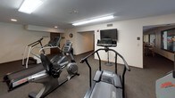 This single-level fitness center is designed to support a well-rounded workout experience within a compact, functional layout. The space is carpeted throughout and illuminated by a combination of recessed lighting and overhead fluorescent fixtures, creating a bright and practical environment for exercise.

The cardio section is equipped with multiple treadmills, elliptical machines, and a stationary bike, with a wall-mounted television positioned for convenient viewing during workouts. A wall clock and motivational fitness posters add character to the space. Strength training is supported by a multi-function cable machine and a dedicated weight bench, providing options for resistance-based exercise.

Full-length mirrors line one wall, visually expanding the room and allowing users to monitor their form. A high-top table with bar-height chairs occupies one corner, offering a rest and refreshment area. Large windows along one side of the room overlook an adjacent indoor pool area, adding visual openness to the space. HVAC vents are positioned throughout the ceiling, ensuring consistent climate control. A solid wood-framed entry door completes the space with a clean, functional aesthetic.