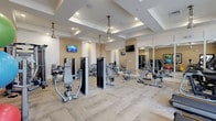 This spacious fitness center is thoughtfully designed to support a comprehensive workout experience. The room is anchored by an elegant coffered ceiling with recessed lighting and pendant-style ceiling fixtures, lending the space a polished, upscale ambiance. Neutral warm-toned walls and light-toned textured carpeting contribute to a clean, welcoming environment throughout.

The facility is outfitted with a robust selection of Matrix-branded equipment, including multiple treadmills and elliptical machines arranged along one wall for cardio training. A dedicated strength training section features an array of selectorized weight machines and cable systems, including a Matrix functional trainer with a full suite of attachments. Benches and additional resistance machines are distributed throughout the floor to accommodate a variety of strength exercises.

Large floor-to-ceiling mirrors line one wall, visually expanding the space and allowing users to monitor their form during workouts. Multiple flat-screen televisions are mounted at strategic points around the room for entertainment during exercise sessions. A functional training corner includes stability balls in multiple sizes, resistance bands, exercise mats, and a medicine ball rack, catering to balance and flexibility training. Natural light filters in through windows and a glass-paneled door, enhancing the overall brightness and energy of the space.
