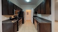 This 1-bedroom, 1-bathroom residence at Hampton Court is thoughtfully laid out across a single floor, offering a comfortable and functional living environment. The galley-style kitchen is well-appointed with dark espresso cabinetry, dark granite-style countertops, stainless steel appliances including a refrigerator, microwave, and dishwasher, plus an electric range. Light tile flooring and crown molding detail add a polished finish to the kitchen space. The living area is generously sized, featuring neutral-toned carpet, a ceiling fan with light fixture, and access to natural light through a large window with outdoor views of lush greenery. The bedroom continues the same warm, carpeted flooring and includes its own ceiling fan, creating a calm and comfortable retreat. A secondary carpeted room provides additional flexible space, suitable for use as a home office or storage area. The bathroom is outfitted with wood-look plank flooring, a tub and shower combination with a curved curtain rod, a dark vanity with granite-style countertop, and clean white fixtures throughout. The overall aesthetic of the unit is clean and neutral, making it a versatile and move-in ready space.