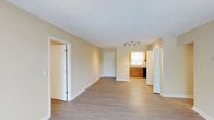 This single-floor space offers a clean, open layout designed for comfortable everyday living. The main living area is spacious and airy, featuring light-toned hardwood-style flooring that runs throughout, contributing to a cohesive and modern aesthetic. Soft, neutral wall tones in warm beige complement the bright white trim, baseboards, and panel-style doors, creating a welcoming and versatile atmosphere.

The open floor plan flows naturally from the living area toward the kitchen, which is visible at the far end of the space. The kitchen features warm wooden cabinetry, a sink beneath a window that allows natural light to filter in, and a dark-finish dishwasher alongside a white refrigerator. A track lighting fixture overhead provides targeted illumination for the kitchen workspace.

Multiple doorways lead to additional rooms, offering a sense of connectivity and thoughtful spatial organization throughout the unit. The layout is practical and uncluttered, making it well-suited for a variety of furnishing arrangements. The smooth white ceiling and consistent flooring throughout help to visually expand the space, while the abundance of natural light enhances the overall bright and inviting character of the interior.