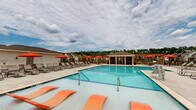 Located in Calabash, North Carolina, this outdoor community amenity area at Brightwater Way offers a well-appointed resort-style retreat designed for relaxation and social gathering. At the heart of the space is a generously sized swimming pool with crystal-clear blue water, framed by an expansive concrete pool deck lined with lounge chairs and vibrant orange umbrellas that provide ample shade throughout the day. A standout architectural feature is the screened-in pavilion situated along the pool's edge, outfitted with comfortable seating and ceiling fans, offering a sheltered yet open-air environment ideal for year-round enjoyment. Adjacent to the pavilion, a wooden pergola with additional umbrella-shaded dining tables and chairs creates a dedicated outdoor gathering space, complete with a grill station for entertaining. The pool deck is enclosed by a wrought-iron style fence, providing security while maintaining clear sightlines to the surrounding residential neighborhood and mature tree line. The community clubhouse anchors the far end of the pool area, featuring a standing-seam metal roof and French doors that open directly onto the deck. Together, these elements create a cohesive, resort-inspired outdoor amenity that serves as a central social hub for the Brightwater Way community.
