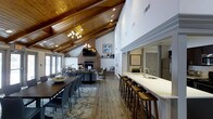 This inviting single-floor space makes an immediate impression with its dramatic vaulted ceilings clad in rich, warm-toned wood planking, supported by exposed beams and illuminated by a statement chandelier. The open-concept layout flows seamlessly from a generous dining area — anchored by a large, dark-wood communal table with ample seating — into a comfortable lounge area furnished with plush sofas and accent chairs arranged around a striking floor-to-ceiling brick fireplace. A mounted television above the fireplace mantel adds a functional focal point to the living area. Natural light pours in through large doors and windows, enhancing the warm, welcoming ambiance throughout. Along one side of the space, a long white quartz-topped bar counter with built-in sink and bar-height wood stools provides an ideal setup for catering or casual refreshments. The durable hardwood-style flooring ties the entire space together, while layered area rugs define the distinct seating zones. Recessed lighting complements the chandelier to ensure the space is well-lit at all hours. The overall atmosphere is refined yet comfortable, making this space well-suited for community gatherings, private events, or social functions.