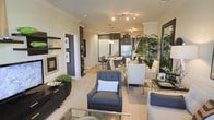 This single-floor residence offers an open-concept layout that effortlessly connects the living, dining, and kitchen areas into one cohesive and well-appointed space. The living area is anchored by plush upholstered seating in deep charcoal tones, complemented by a sleek glass coffee table that reflects the warm, ambient lighting overhead. Crown molding runs throughout, adding a refined architectural detail that elevates the overall aesthetic.

The dining area transitions naturally from the living space, featuring a clean-lined table surrounded by crisp white chairs with chrome accents, creating a modern yet inviting atmosphere. Lush indoor plants add a vibrant, organic touch that softens the contemporary design palette.

The kitchen is defined by dark cabinetry contrasted against lighter walls, with stainless steel appliances and stylish pendant lighting suspended above the counter. Track lighting throughout ensures the space is well-lit and visually dynamic.

A hallway leads to additional areas of the home, framed by white paneled doors and warm wood-toned flooring. Decorative wall art and carefully curated accessories are displayed throughout, reflecting an attention to detail and a cohesive design vision. The overall ambiance is sophisticated, warm, and thoughtfully designed for both relaxed living and entertaining.