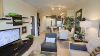This single-floor residence offers an open-concept layout that effortlessly connects the living, dining, and kitchen areas into one cohesive and well-appointed space. The living area is anchored by plush upholstered seating in deep charcoal tones, complemented by a sleek glass coffee table that reflects the warm, ambient lighting overhead. Crown molding runs throughout, adding a refined architectural detail that elevates the overall aesthetic.

The dining area transitions naturally from the living space, featuring a clean-lined table surrounded by crisp white chairs with chrome accents, creating a modern yet inviting atmosphere. Lush indoor plants add a vibrant, organic touch that softens the contemporary design palette.

The kitchen is defined by dark cabinetry contrasted against lighter walls, with stainless steel appliances and stylish pendant lighting suspended above the counter. Track lighting throughout ensures the space is well-lit and visually dynamic.

A hallway leads to additional areas of the home, framed by white paneled doors and warm wood-toned flooring. Decorative wall art and carefully curated accessories are displayed throughout, reflecting an attention to detail and a cohesive design vision. The overall ambiance is sophisticated, warm, and thoughtfully designed for both relaxed living and entertaining.
