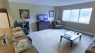 This single-floor residence offers a comfortable and functional layout designed to maximize livability. The open-concept living area is a central highlight, featuring soft wall-to-wall carpeting that adds warmth underfoot and a neutral color palette on the walls that creates a calm, inviting atmosphere. A large window floods the living space with natural light and provides pleasant views of the surrounding greenery, enhancing the sense of openness throughout the room. The living area is well-proportioned, comfortably accommodating a seating arrangement alongside a media console, making it an ideal space for both relaxing and entertaining. Framed artwork on the walls adds a touch of personality and visual interest. Transitioning seamlessly from the living area, the dining space is positioned along a granite-topped counter or bar area, offering a practical surface for meals or casual gathering. The dining setup is neatly arranged with place settings, suggesting a thoughtful approach to both style and function. A baseboard heating system is integrated along the walls, ensuring year-round comfort. Overall, the space presents a clean, well-maintained interior that balances everyday practicality with a welcoming residential ambiance.