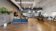 Located at 612 W Main Ave in Gastonia, NC, this single-floor commercial space blends industrial character with modern, thoughtfully curated design. Rich hardwood floors run throughout, complementing the exposed structural columns, overhead ductwork, and track lighting that lend an authentic urban loft aesthetic.

The open-plan layout is divided into distinct functional zones. A lounge area features tufted leather sofas, a solid wood coffee table, and a large flat-screen television mounted against a striking accent wall adorned with an angular geometric pattern of raw timber beams — a true focal point of the space. Decorative shelving, framed artwork, and potted fiddle-leaf fig trees add warmth and personality.

A recreation area includes a billiards table beneath stylish pendant lighting, providing an inviting space for leisure and social interaction. Adjacent to this is a well-appointed kitchenette, lined with sleek gray cabinetry, stainless steel appliances, and counter seating. The defining feature here is a dramatic floor-to-ceiling typographic mural filled with motivational phrases, set against a dark background for bold visual impact.

A reception-style nook near the entrance offers a workstation desk and comfortable seating alongside glass-paneled entry doors. Throughout, the steel-blue wall palette, exposed infrastructure, and curated décor create a cohesive, energetic, and welcoming environment.