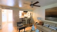This single-floor residence offers a welcoming and functional layout designed for comfortable everyday living. The living area serves as the heart of the home, featuring warm hardwood-style flooring, neutral-toned walls, and a ceiling fan with integrated lighting that provides both style and practical air circulation. The space is furnished with a plush sofa, a cushioned accent chair, and a glass coffee table adorned with decorative vases, creating an inviting and well-curated atmosphere.

A defining architectural feature is the open pass-through window that seamlessly connects the living room to the kitchen, enhancing the sense of openness and allowing natural light from the kitchen window to filter through. The kitchen is equipped with dark wood cabinetry, a refrigerator, and a range hood, and opens to a breakfast bar fitted with tall bar stools — ideal for casual dining or entertaining.

The overall ambiance is warm and understated, with soft ambient lighting from table lamps complementing the natural light. The cohesive color palette of taupes, grays, and warm wood tones ties the living and kitchen spaces together, making this a practical and aesthetically pleasing single-floor home.