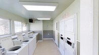 This single-floor laundry facility is designed with practicality and efficiency in mind. The room is finished with soft green walls accented by a gray wainscoting panel along the lower half, creating a clean and organized aesthetic throughout the space. Bright flush-mounted fluorescent light panels in the ceiling ensure excellent visibility at all times of day, while a window along one wall allows for natural light to supplement the interior lighting.

Along one side of the room, a row of top-load washing machines is neatly arranged, providing ample capacity for multiple users. On the opposite side, stacked washer and dryer units with coin-operated controls offer additional washing and drying options, maximizing the use of available space. The units are all white, contributing to the room's bright and tidy appearance.

The flooring is a light-toned, easy-to-clean surface well-suited for a high-use utility space. A wall-mounted ventilation unit helps maintain air quality and manage humidity levels within the room. Posted signage provides clear instructions and guidelines for users. A doorway at the far end of the room offers access to an adjoining area. Overall, this laundry facility is a functional, well-maintained space built to serve a shared community efficiently.