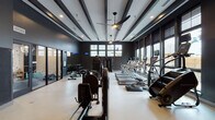 This fitness center presents a thoughtfully designed workout environment anchored by a striking dark color palette. Deep charcoal walls contrast with light-toned flooring, creating a bold, contemporary aesthetic throughout the space. The ceiling features distinctive exposed beam detailing finished in dark tones, complemented by ceiling fans and recessed lighting that maintain a comfortable atmosphere during workouts.

Natural light flows generously through large, dark-framed windows lining the walls, offering exterior views while keeping the interior bright and energizing. The main workout area is generously proportioned, providing ample room between equipment stations. A row of treadmills and stair climbers with individual screens lines the window-facing wall, ideal for cardio training with an outward view.

The strength training section includes cable machines, a multi-function gym station, and a well-stocked dumbbell rack accompanied by a flat bench, all positioned in front of floor-to-ceiling mirrors with digital clocks for tracking workouts. Additional equipment such as rowing machines and cable systems are thoughtfully distributed throughout the floor.

A separate enclosed room, accessible through glass-paneled doors, provides a dedicated area for additional training activities. A wall-mounted television and clean entry corridor complete this polished, fully equipped facility.