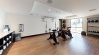 This single-floor fitness studio is designed to support a comprehensive workout experience within a clean, contemporary setting. The space is finished with warm hardwood-style flooring and crisp white walls accented by dark wood baseboards, creating a polished and inviting atmosphere throughout.

The main exercise area is generously proportioned, accommodating multiple stations without feeling crowded. A row of spin bikes serves as a centerpiece for cycling workouts, while a ceiling-mounted TRX suspension training rig offers functional strength and flexibility training options. Along one wall, a dedicated rack holds an organized selection of kettlebells in varying weights alongside medicine balls, ensuring versatility for strength-based routines. Additional medicine balls are mounted on the wall for easy access.

Natural light enters through a large floor-to-ceiling window, offering an outdoor view and brightening the workout environment. A wall-mounted Fitness On Demand screen near the entrance provides guided, on-demand class programming, adding an interactive dimension to the space.

The studio is accessed through dark-framed glass double doors, and recessed ceiling lighting ensures consistent illumination throughout. Decorative wall art adds a subtle creative touch, contributing to a motivating and well-appointed fitness environment.