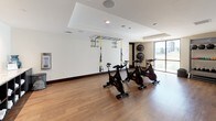 This single-floor fitness studio is designed to support a comprehensive workout experience within a clean, contemporary setting. The space is finished with warm hardwood-style flooring and crisp white walls accented by dark wood baseboards, creating a polished and inviting atmosphere throughout.

The main exercise area is generously proportioned, accommodating multiple stations without feeling crowded. A row of spin bikes serves as a centerpiece for cycling workouts, while a ceiling-mounted TRX suspension training rig offers functional strength and flexibility training options. Along one wall, a dedicated rack holds an organized selection of kettlebells in varying weights alongside medicine balls, ensuring versatility for strength-based routines. Additional medicine balls are mounted on the wall for easy access.

Natural light enters through a large floor-to-ceiling window, offering an outdoor view and brightening the workout environment. A wall-mounted Fitness On Demand screen near the entrance provides guided, on-demand class programming, adding an interactive dimension to the space.

The studio is accessed through dark-framed glass double doors, and recessed ceiling lighting ensures consistent illumination throughout. Decorative wall art adds a subtle creative touch, contributing to a motivating and well-appointed fitness environment.