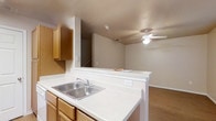 This two-story residence offers a functional and welcoming layout designed for comfortable everyday living. The main level features an open-concept design that seamlessly connects the kitchen and living area, creating a spacious and cohesive environment. The kitchen is equipped with warm honey-toned wood cabinetry that provides ample storage, complemented by a white countertop peninsula that doubles as a breakfast bar or casual dining surface. A stainless steel double-basin sink is positioned along the peninsula, offering a practical workspace with a view into the adjoining living area. The living space is outfitted with a ceiling fan, promoting comfortable airflow throughout the room. Durable hardwood-style flooring runs throughout the main level, adding warmth and easy maintenance to the space. A staircase is visible from the main living area, providing access to the upper floor. The neutral color palette of warm whites and soft beiges throughout the space creates a clean, airy ambiance that serves as a versatile backdrop. The open sightlines between the kitchen and living area make this home well-suited for both everyday living and entertaining.