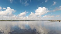 This single-level space is defined by its remarkable waterfront setting, offering unobstructed panoramic views of a broad, calm body of water that stretches to a distant tree-lined horizon. The water's surface mirrors the sky above, creating a serene and visually expansive atmosphere. Billowing cumulus clouds drift across a vivid blue sky, enhancing the sense of openness and natural beauty that characterizes this space.

The outdoor vantage points provide a sweeping perspective across the water, with lush, low-lying greenery visible along the far shoreline. The tranquil and reflective quality of the water contributes to a peaceful ambiance throughout. The surrounding landscape is flat and open, typical of a coastal or estuarine environment, allowing for uninterrupted sightlines in multiple directions.

Interior spaces within the structure are present, though they are minimally illuminated in the available imagery, revealing only subtle architectural details such as doorways, fixtures, and interior surfaces. The overall character of this space is shaped primarily by its exceptional connection to the natural waterfront environment, making the outdoor views and proximity to the water the defining features of the property.