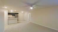 This single-floor space features an open-concept layout that seamlessly connects the living area and kitchen. The living area is finished with light-toned carpeting and neutral cream-colored walls, creating a bright and understated atmosphere. A ceiling fan with a built-in light fixture is centrally positioned overhead, providing both airflow and ambient lighting, complemented by recessed lighting throughout.

The kitchen is visibly integrated into the space, featuring dark cabinetry that provides a contrasting accent against the lighter surroundings. A stainless steel refrigerator anchors the kitchen, and a peninsular counter separates it from the main living area, offering a natural transition between the two zones. The dark upper cabinets add a modern touch to the overall neutral palette.

Two doorways are visible from the living area, suggesting access to additional rooms or storage spaces within the unit. White baseboards line the perimeter of the space, adding clean definition to the flooring. The overall layout is open and versatile, offering a functional and move-in-ready environment suitable for a variety of furniture arrangements and personal styles.