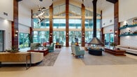 Located at 4961 Woodruff Springs Rd in Sanford, Florida, this single-story clubhouse at Integra Leasing is a thoughtfully designed community amenity space that blends contemporary architecture with vibrant, curated interiors. The grand entrance foyer sets an impressive tone, featuring soaring vaulted ceilings, floor-to-ceiling glass panels framed by rich wood columns, and sculptural pendant lighting that draws the eye upward. Views of lush palm trees beyond the expansive glazing reinforce a connection to the Florida landscape.

The main lounge centers around a striking suspended conical fireplace, surrounded by mid-century modern seating in teal, cream, and warm wood tones. Adjacent areas offer flexible social spaces with distinct design identities, including a cozy lounge with tray ceilings, ambient lighting, and plush sectional seating, and a bold accent room showcasing textured wood-panel walls and teal wainscoting.

A well-appointed social kitchen and dining area features a large white quartz island with bar seating, statement globe pendant fixtures, a patterned tile floor, and a wall-mounted TV. The game room extends the entertainment offerings with a pool table, a kitchenette, and direct outdoor access. Glass-walled conference rooms provide functional workspace within the clubhouse footprint.