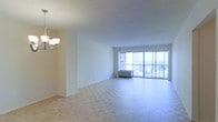 This single-floor space offers an open and airy layout centered around a generously sized living area. The room is finished with classic parquet hardwood flooring arranged in a herringbone-style pattern, adding warmth and texture to the bright interior. Neutral white walls keep the space feeling clean and versatile, ready to accommodate a range of furnishings and design styles.

A standout feature of the living area is the expansive floor-to-ceiling window wall that floods the interior with abundant natural light throughout the day. A sliding glass door provides direct access to a private balcony, where panoramic views of the surrounding cityscape and distant landscape can be enjoyed. The outdoor setting visible through the glass highlights an elevated position within a high-rise building.

Overhead, a multi-arm chandelier provides ambient lighting to complement the natural light. A wall-mounted climate control unit near the window ensures year-round comfort. The overall ambiance is clean, bright, and open, making this a functional and inviting living space with a strong connection to its urban surroundings.
