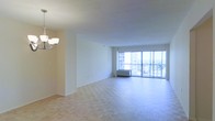 This single-floor space offers an open and airy layout centered around a generously sized living area. The room is finished with classic parquet hardwood flooring arranged in a herringbone-style pattern, adding warmth and texture to the bright interior. Neutral white walls keep the space feeling clean and versatile, ready to accommodate a range of furnishings and design styles.

A standout feature of the living area is the expansive floor-to-ceiling window wall that floods the interior with abundant natural light throughout the day. A sliding glass door provides direct access to a private balcony, where panoramic views of the surrounding cityscape and distant landscape can be enjoyed. The outdoor setting visible through the glass highlights an elevated position within a high-rise building.

Overhead, a multi-arm chandelier provides ambient lighting to complement the natural light. A wall-mounted climate control unit near the window ensures year-round comfort. The overall ambiance is clean, bright, and open, making this a functional and inviting living space with a strong connection to its urban surroundings.