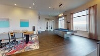 This dynamic single-floor amenity space is thoughtfully designed to foster social interaction and recreation. Upon entry, visitors are welcomed by a bright foyer featuring rich wood-look flooring, a bold red pendant light, a curated art piece, and a compact bistro seating nook — setting a contemporary and inviting tone throughout.

The space flows into a generously sized game and lounge area, where dark hardwood flooring anchors an eclectic mix of furnishings and activities. A sleek pool table with a blue felt top serves as a centerpiece, accompanied by a foosball table, a classic arcade machine, and a wall-mounted television. Comfortable lounge seating, a communal dining table, and ambient cove lighting overhead create a relaxed yet energetic atmosphere.

A dedicated lounge alcove features a wall-mounted wooden counter with colorful bar stools and vibrant abstract wall art. This area opens directly through floor-to-ceiling black-framed glass doors onto an outdoor patio, where string lights, patio seating, a ping-pong table, and surrounding stone architectural details create an inviting extension of the interior.

Expansive windows throughout flood the space with natural light and frame views of the surrounding landscape. The combination of bold artwork, mixed-material furnishings, and well-considered lighting makes this a versatile and engaging communal destination.